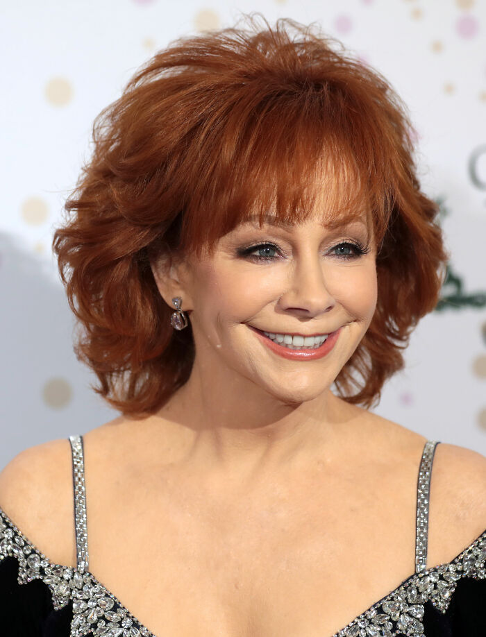 Reba McEntire smiling at an event, wearing a black dress with silver embellishments and styled red hair.