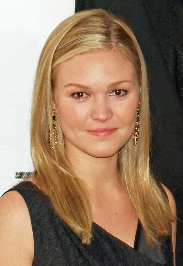 Julia Stiles with straight blonde hair wearing a black dress and gold earrings at a formal event.