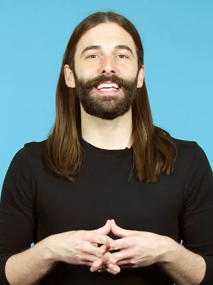 Jonathan Van Ness smiling with long hair and beard, wearing a black shirt against a blue background in a casual pose.