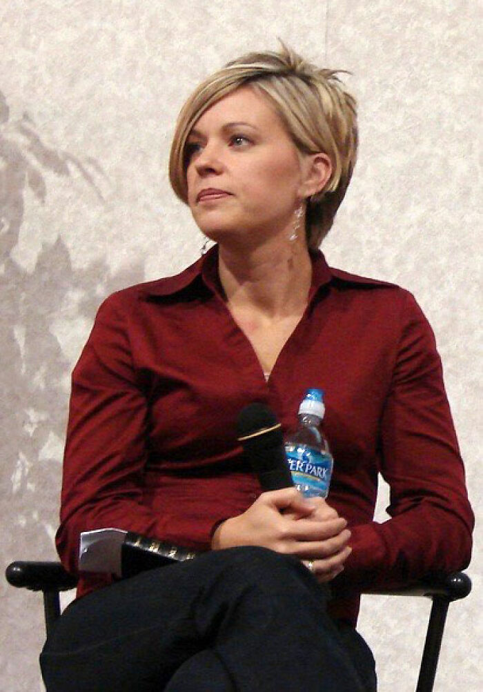 Kate Gosselin sitting on a chair, holding a microphone and water bottle, wearing a red shirt and earrings.