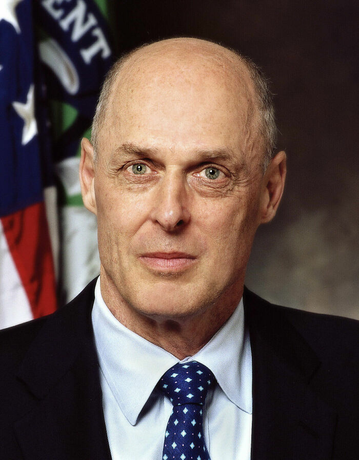 Henry Paulson in formal attire posing for an official portrait with a flag in the background.