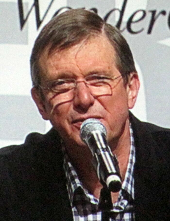 Mike Newell speaking into a microphone at an event, wearing glasses and a checkered shirt under a dark jacket.