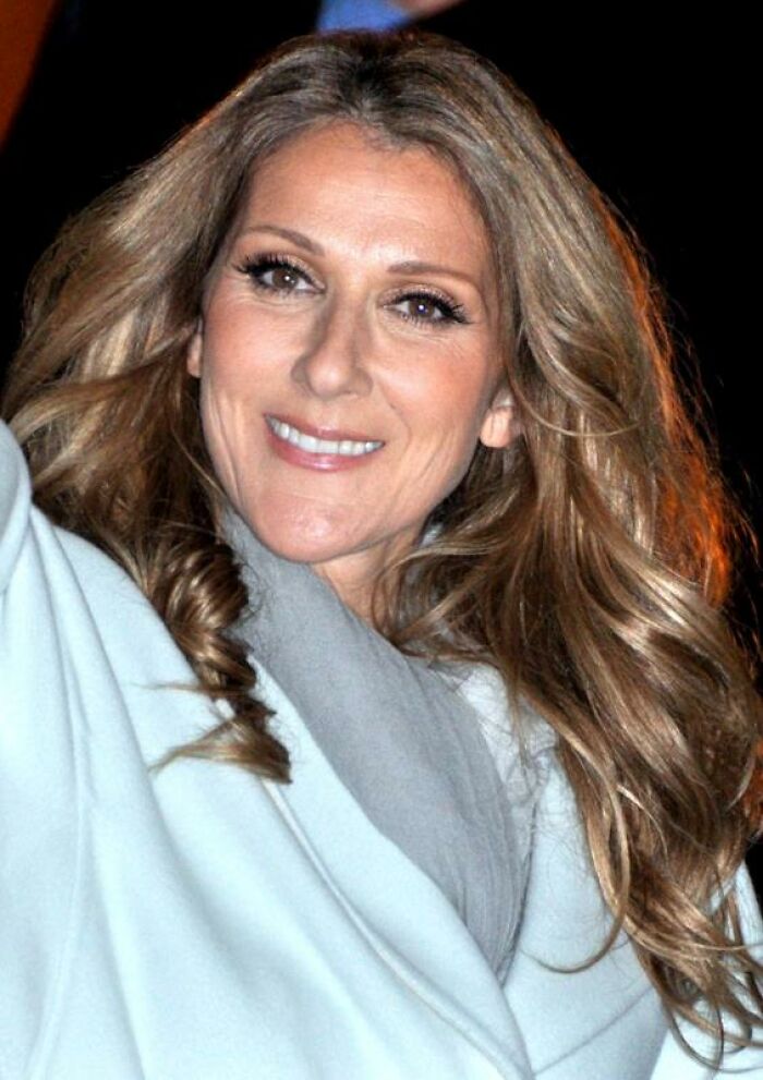 Celine Dion smiling with wavy hair, wearing a light coat and scarf, showcasing her bio and career highlights.