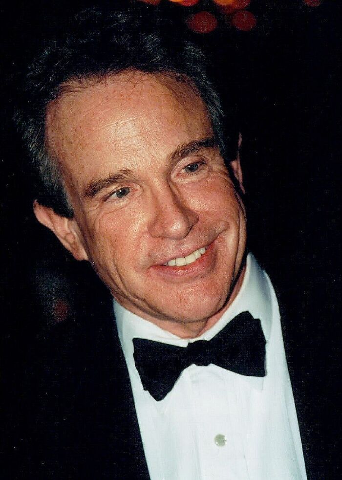 Warren Beatty smiling, wearing a black tuxedo and bow tie at a formal event with dim lighting in the background