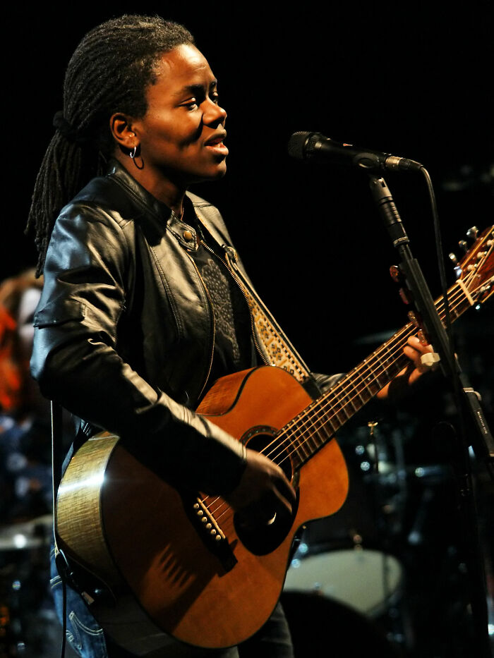 Tracy Chapman performing live on stage, playing acoustic guitar and singing into a microphone during a concert.