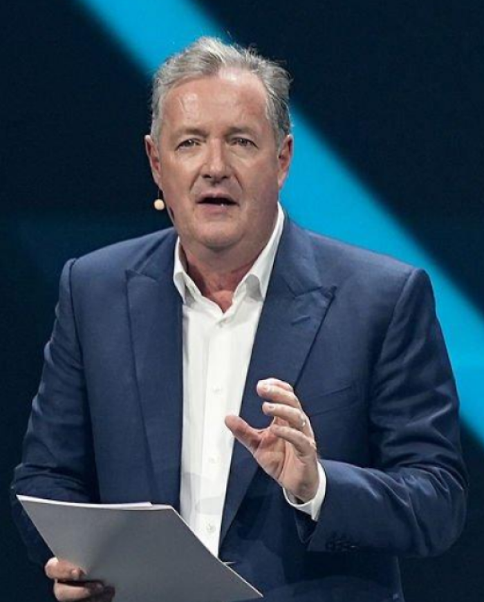 Piers Morgan speaking on stage, wearing a navy suit and white shirt, holding papers during a public event.