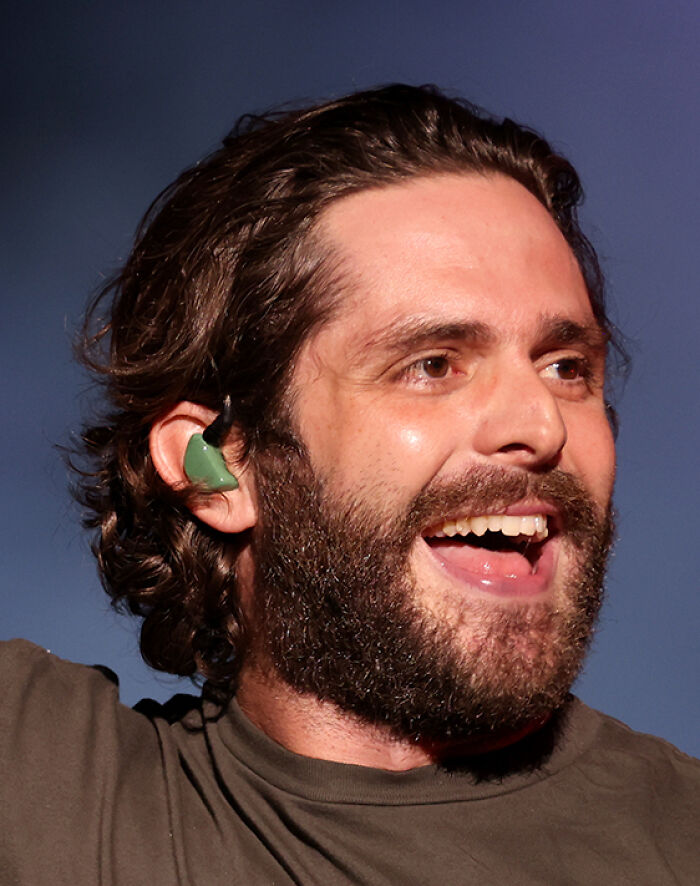 Thomas Rhett smiling during a live performance, wearing a green earpiece and casual dark shirt on stage.