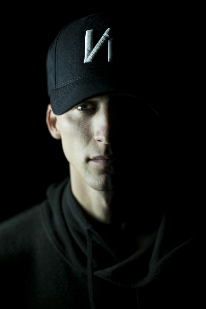 Close-up of a man wearing a black cap with NF logo, half of his face illuminated, representing NF bio and career highlights.