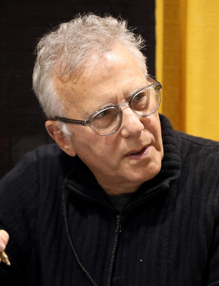 Paul Reiser wearing glasses and a dark sweater, engaging in conversation at an indoor event.