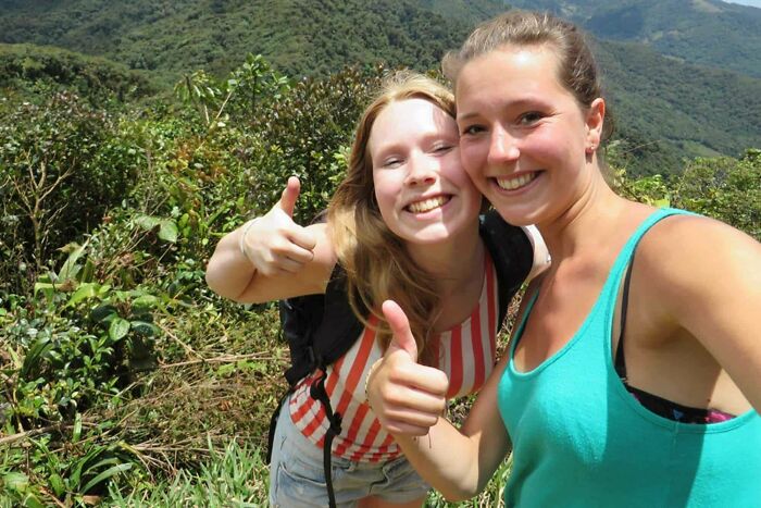Two young women hiking in a lush forest, smiling and giving thumbs up, symbolizing unsettling mysteries in nature.
