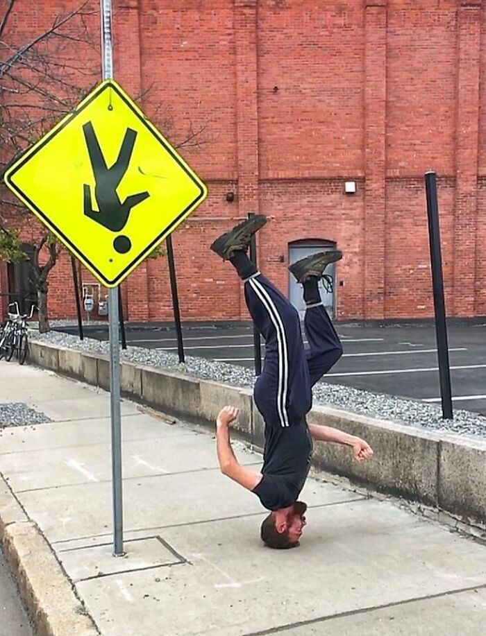 Man pulling off a creative pose by balancing upside down on his head near a funny upside down pedestrian sign.