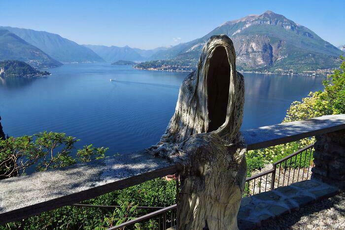 Hooded creepy sculpture with a hollow face overlooking a serene lake and mountains, evoking a weird and eerie vibe.