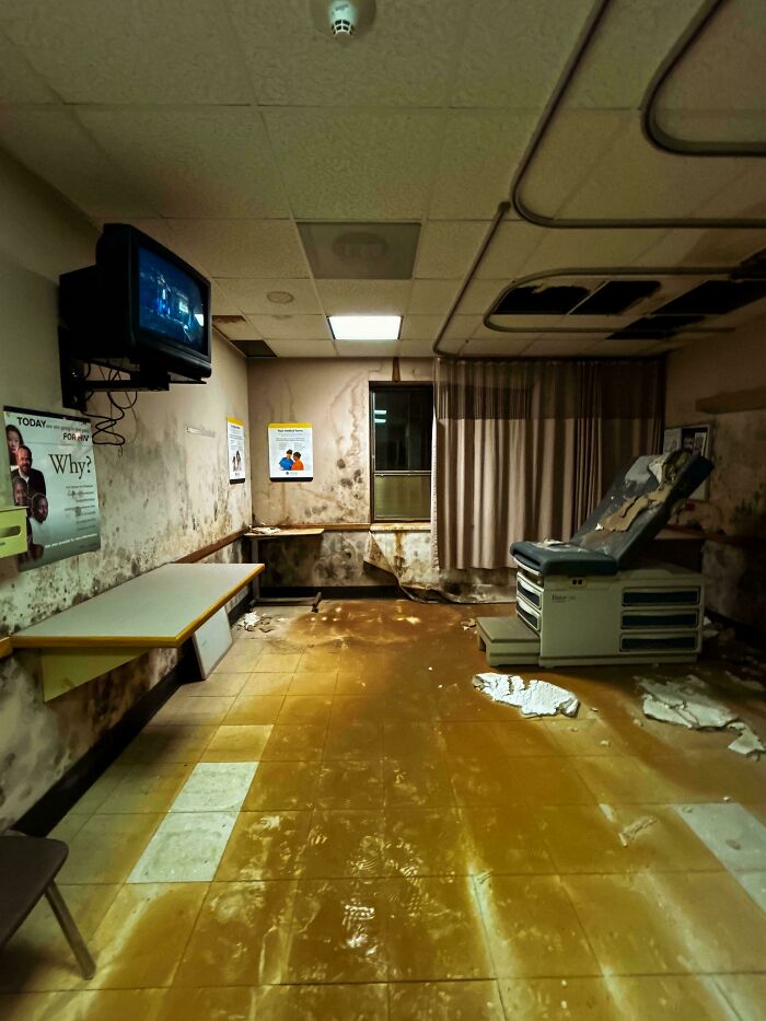 Deteriorated and creepy abandoned medical exam room with mold, peeling walls, and damaged floor in dim lighting.