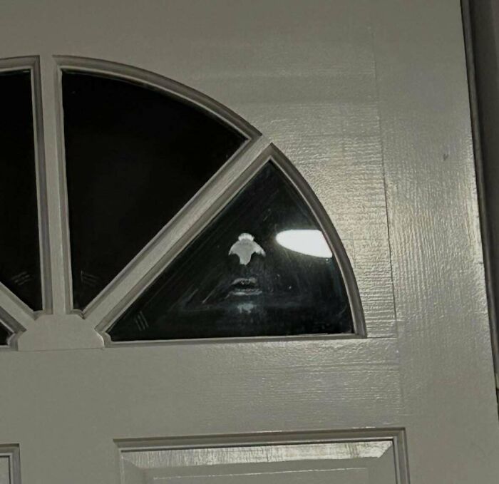 Shadowy face with glowing eyes and eerie mouth behind a door window, creating creepy and weird photo vibes.