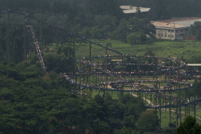 A large crowd walks along a winding roller coaster track in a green, forested area, creating an unsettling mystery scene.