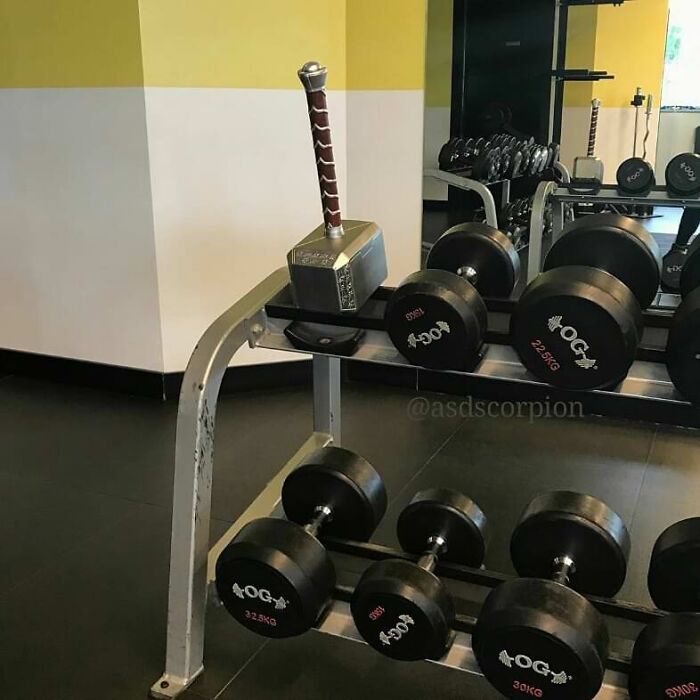 Thor's hammer placed among gym dumbbells on a weight rack, a surprising Darwin Award contender moment.