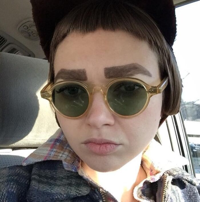 Person with crazy eyebrows drawn thick and square-shaped, wearing round sunglasses and a dark hat inside a car.