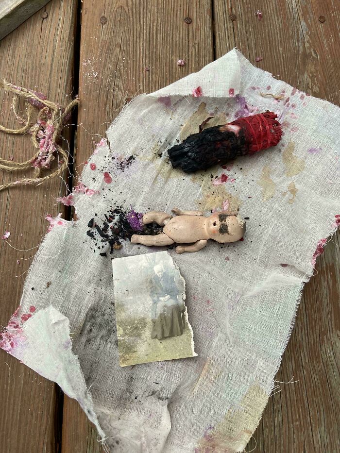 Worn doll and burnt red sage stick on stained cloth with old black and white photograph on wooden surface, unsettling mysteries.
