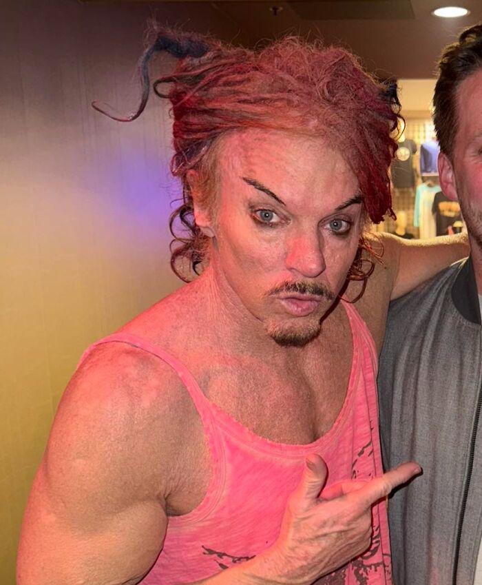 Man with crazy eyebrows styled aggressively, wearing a pink tank top and striking a dynamic pose indoors.