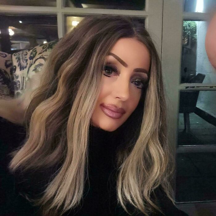 Woman with thick, well-defined crazy eyebrows styled to stand out against her long wavy blonde hair and dark top indoors.