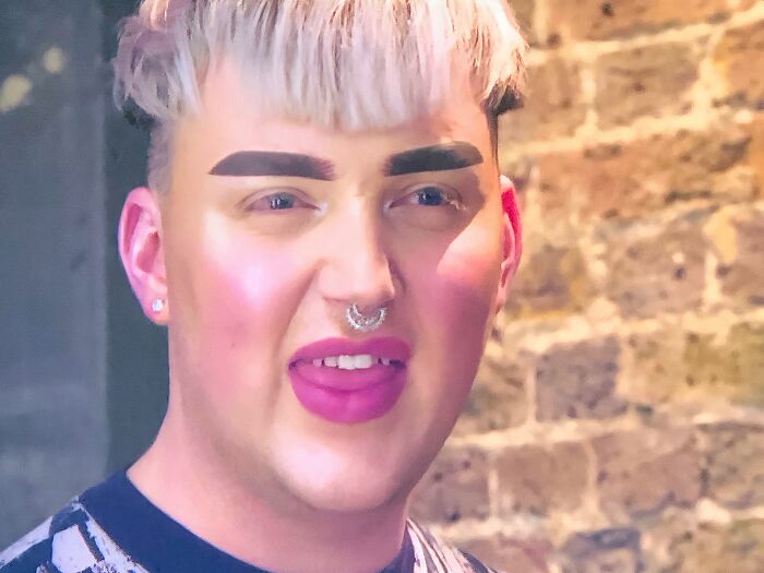 Person with crazy eyebrows styled thick and dark against a brick wall background, wearing bright lipstick and a septum piercing.