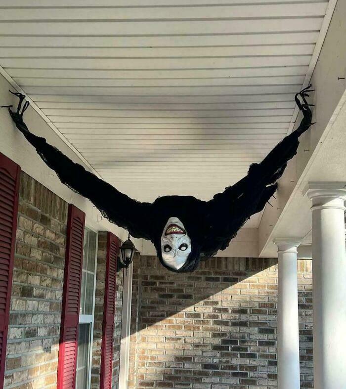 Creepy and weird photo of a hanging ghoul figure with a white mask and long black limbs on a porch ceiling.