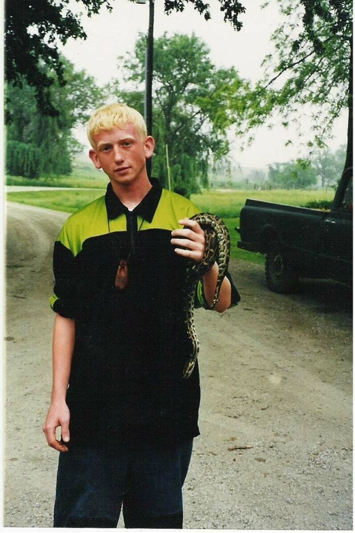 Young man with blond hair holding a snake outdoors on a rural dirt road, evoking unsettling mysteries intrigue.