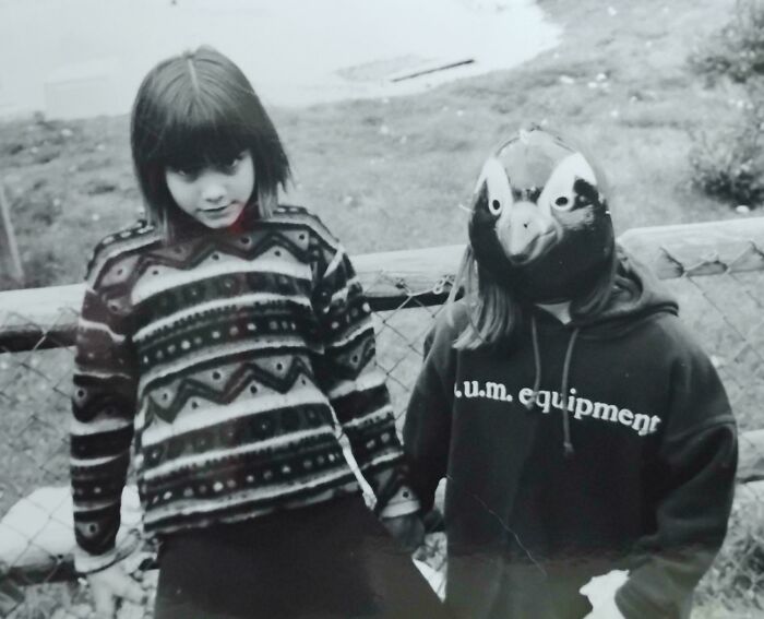 Two children pose by a fence, one wearing a bird mask, creating a creepy and weird photo vibe.