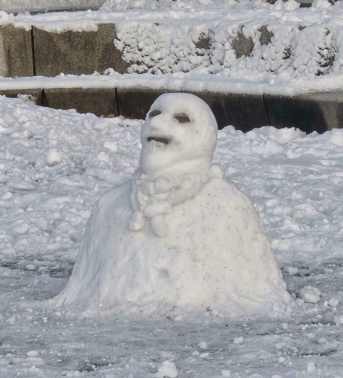 Creepy snow sculpture with eerie face and hollow eyes in a snowy urban setting, evoking weird and creepy vibes.