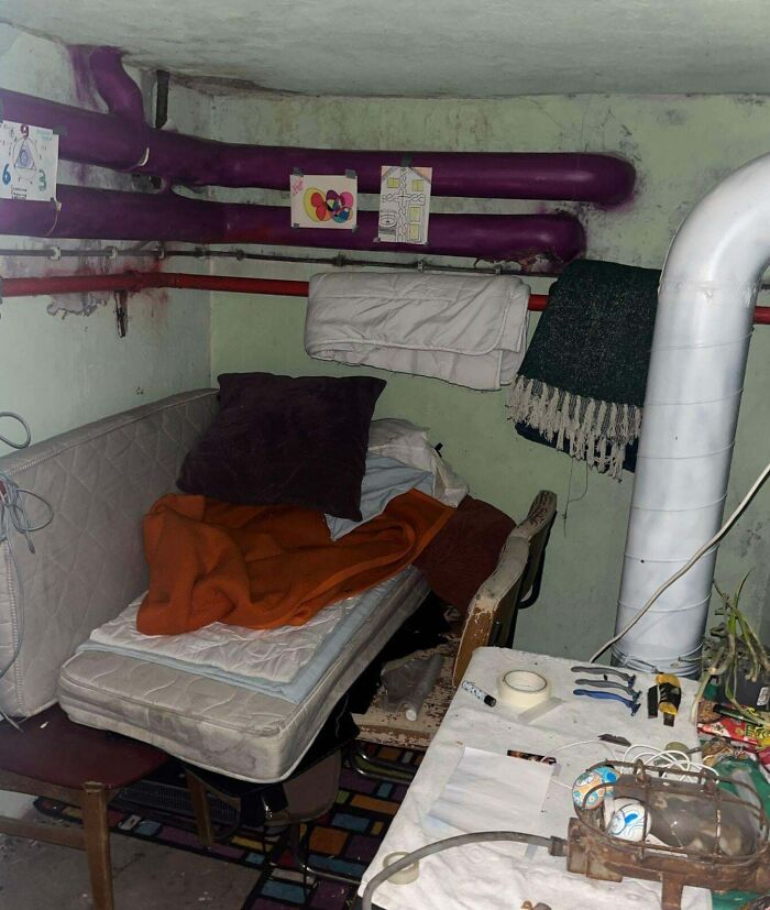 Cluttered, creepy basement room with old mattress, blankets, purple pipes, and a table full of tools and miscellaneous items.