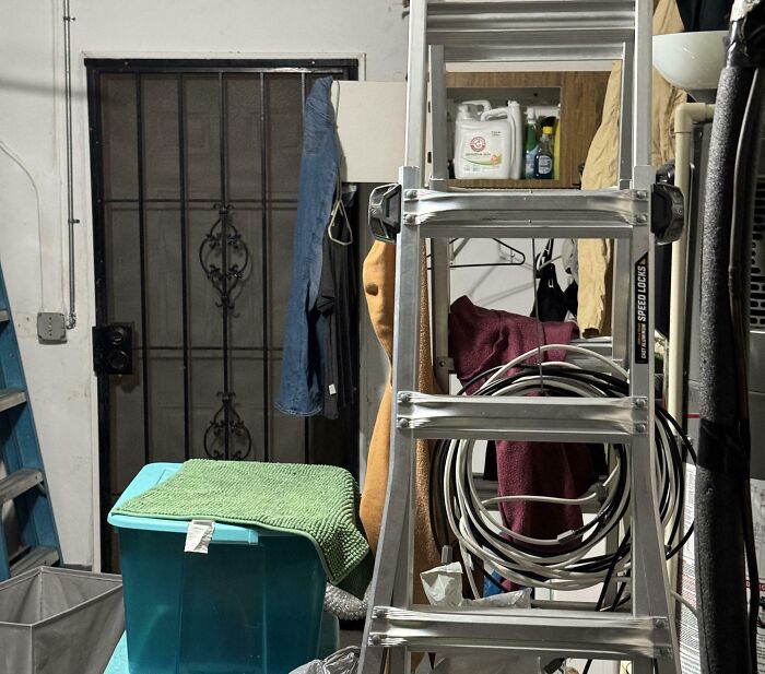 Cluttered storage room with ladder, hanging clothes, coiled cables, and a green plastic bin, creating a creepy and weird atmosphere.