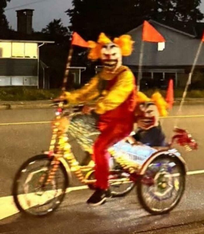 Creepy clown costume with bright orange hair riding a decorated tricycle on a dimly lit street at night.