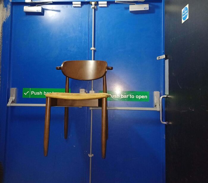 Wooden chair mysteriously suspended in front of blue emergency exit door, creating a creepy and weird visual effect.