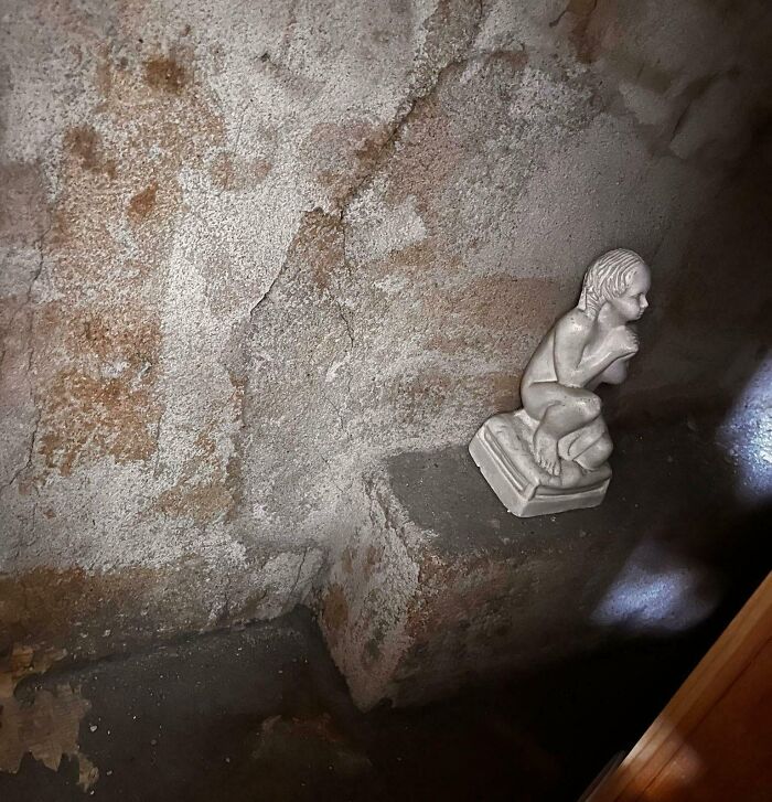 Small creepy statue on a concrete ledge against a cracked wall, evoking eerie and weird vibes in dim lighting.