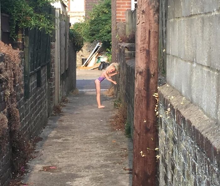 Child bending backward in narrow alleyway between brick and stone walls, creating a creepy and weird photo scene.