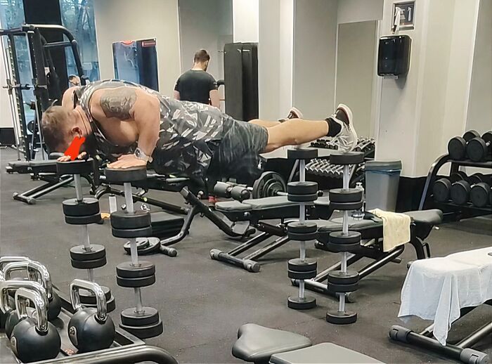 Man attempting a risky plank balancing on dumbbells with feet elevated on a bench in a gym Darwin Award contender moment