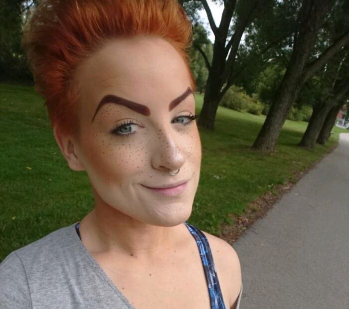 Person with crazy eyebrows shaped in a bold, angular style standing outdoors by a tree-lined path.