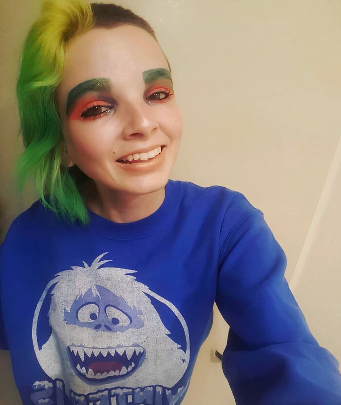 Person with green and yellow hair and crazy eyebrows styled boldly, wearing a blue sweatshirt with a cartoon character.