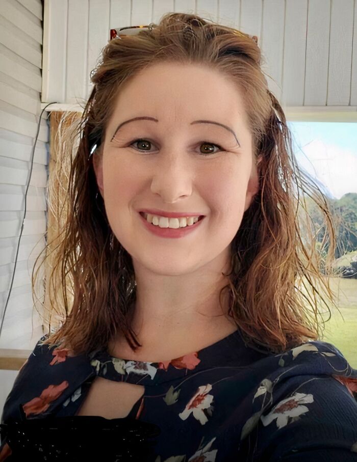 Woman smiling with drawn-on crazy eyebrows that seem to live their own lives on her face indoors by a window.