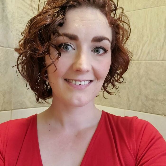 Woman with curly red hair and crazy eyebrows seeming to live their own lives, smiling in a red top indoors.