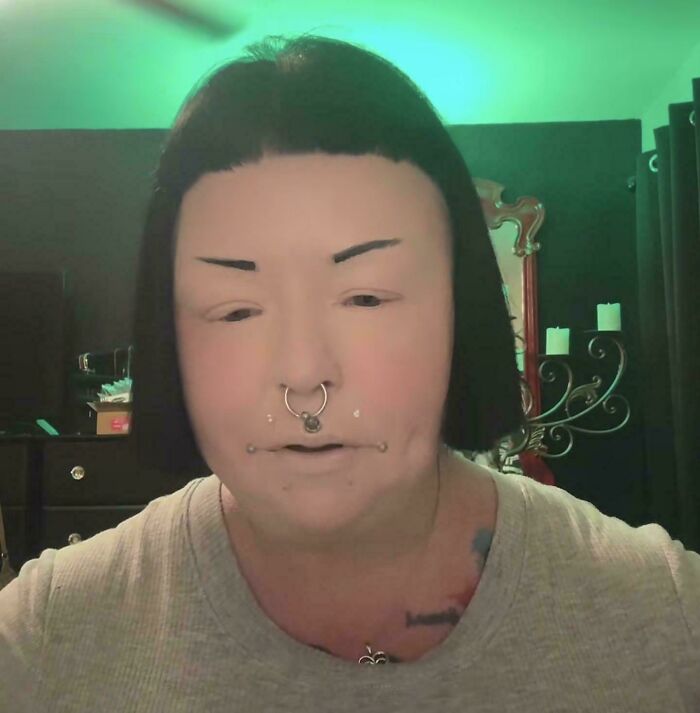 Person with straight, exaggerated crazy eyebrows and multiple facial piercings in a softly lit room with dark decor.