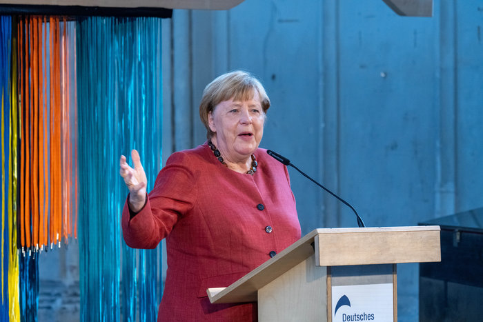Angela Merkel in a red jacket speaking at a podium, representing influential leaders who changed history.