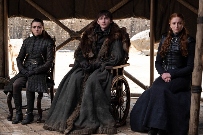 Arya, Bran, and Sansa Stark seated in a tent, dressed in dark medieval attire from the Game of Thrones series.