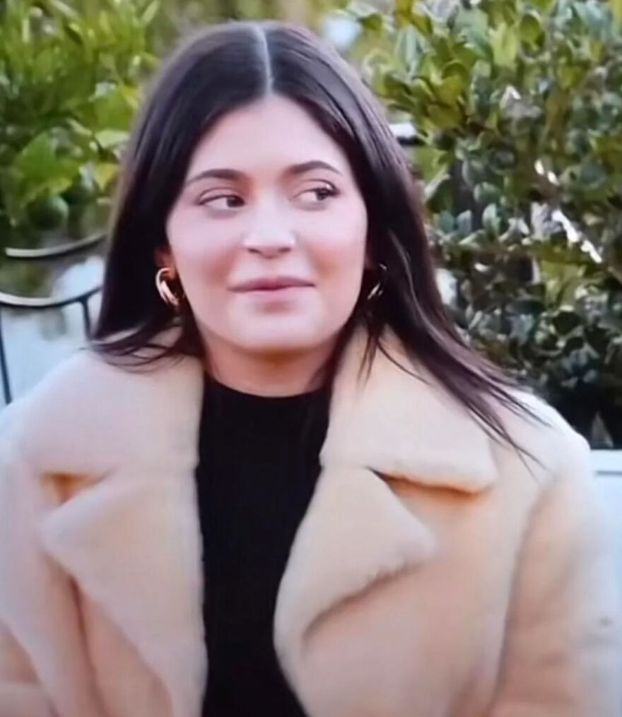 Kylie Jenner up close outdoors wearing a beige fur coat and gold hoop earrings with natural makeup and long dark hair.