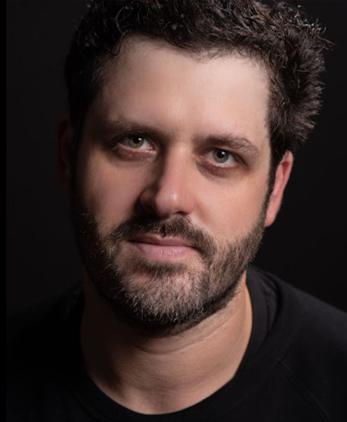 Retrato em close de um homem com cabelo e barba escuros contra um fundo preto relacionado a Adam Pearson lançando um tweet ofensivo.