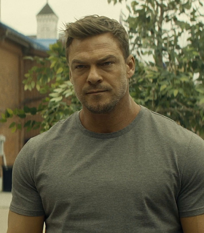 Alan Ritchson outdoors wearing a grey t-shirt, facing forward with a serious expression, greenery in the background