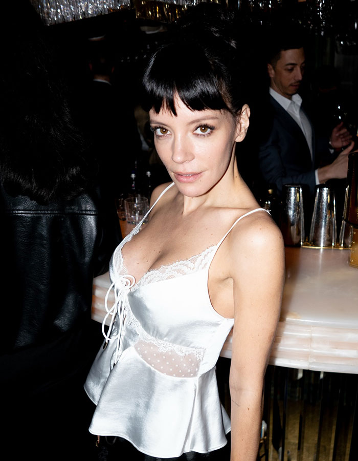 Lily Allen em lingerie branca, com destaque para a cirurgia plástica, posando com confiança em um evento noturno interno.