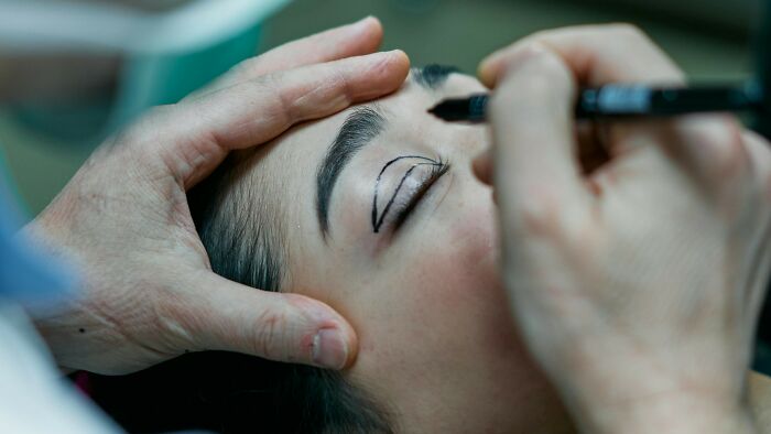 Close-up of a woman undergoing plastic surgery preparation with markings on her eyelids, addressing peer pressure issues.