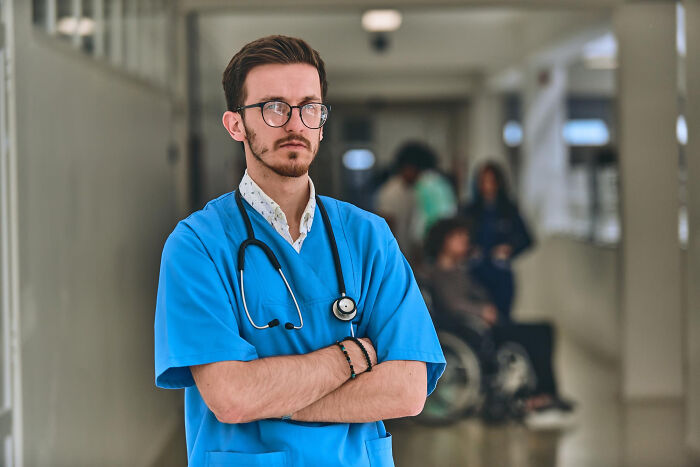 Young male doctor in blue scrubs with stethoscope, standing confidently in hospital hallway with patients in background