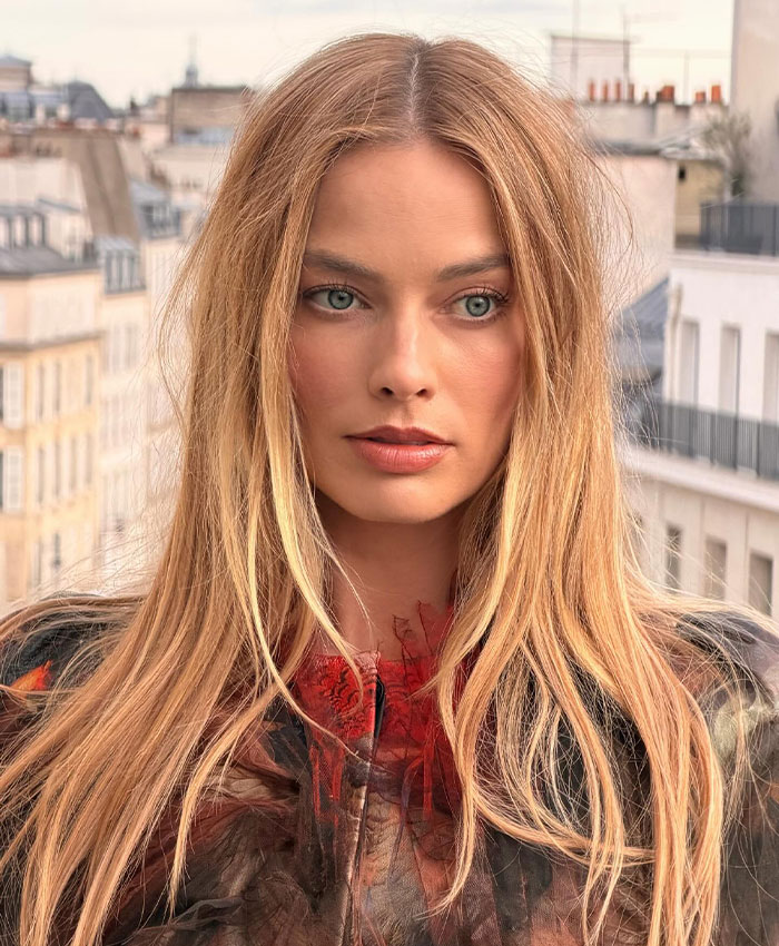 Margot Robbie at Paris Fashion Week, posing with long blonde hair and a sheer red and black outfit.